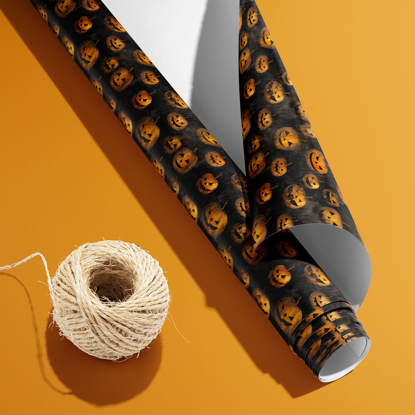 A partially unfurled Jack-o-Lanterns roll with white underside sits by a spool of natural twine, showing glowing pumpkins on smoky black, set on an orange background.