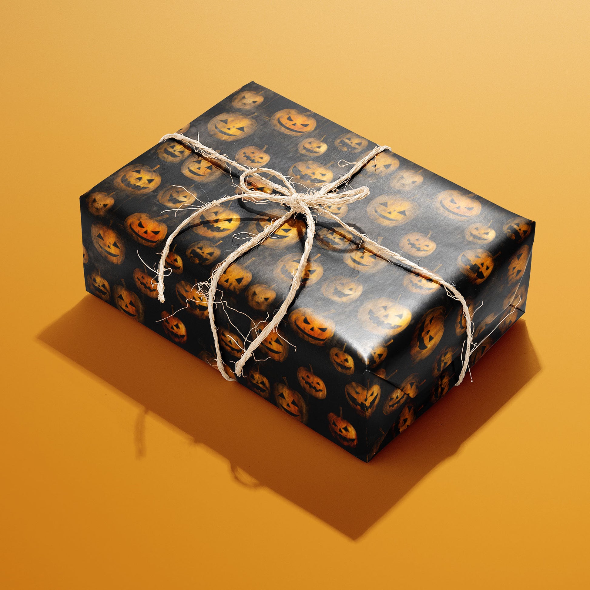 A rectangular gift wrapped in Jack-o-Lanterns paper and tied with natural twine displays glowing carved pumpkins on a smoky black ground against an orange background.
