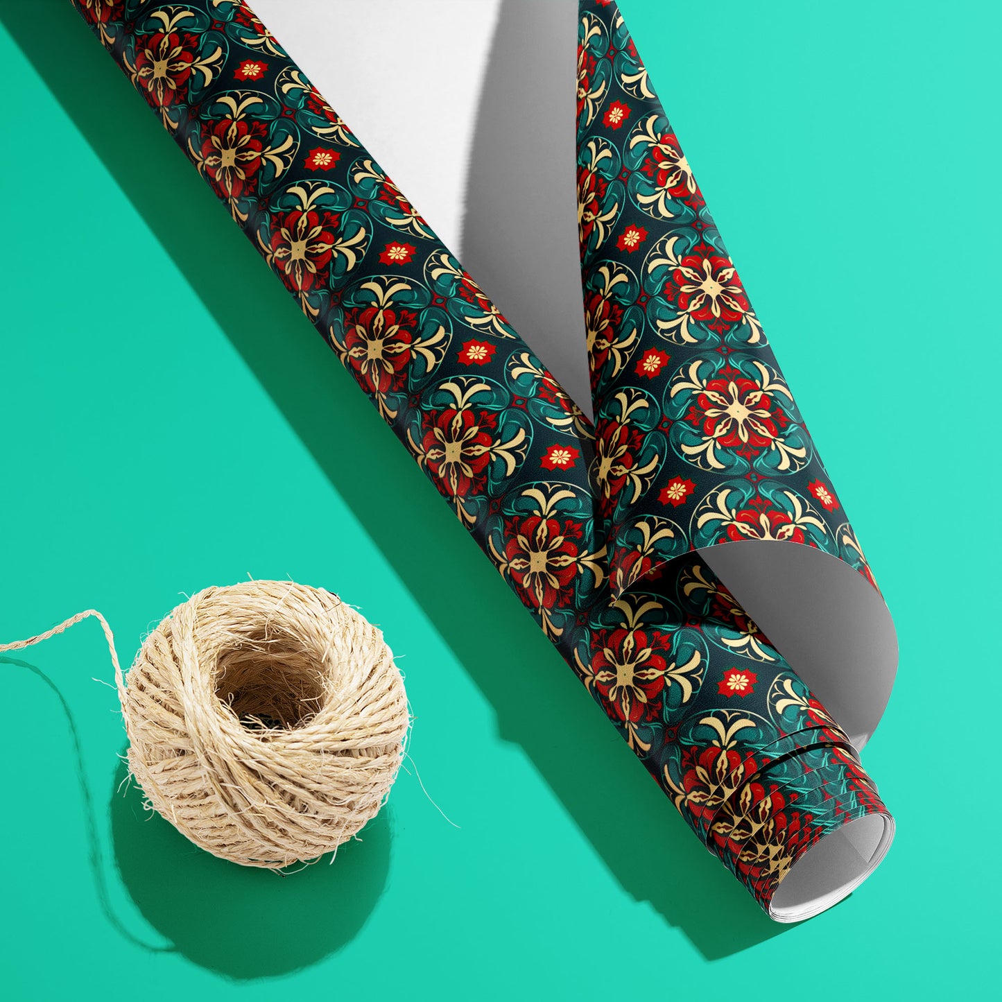 A partially unfurled roll of Arabesque Christmas paper with the white underside showing beside a spool of natural twine on a bright teal background.