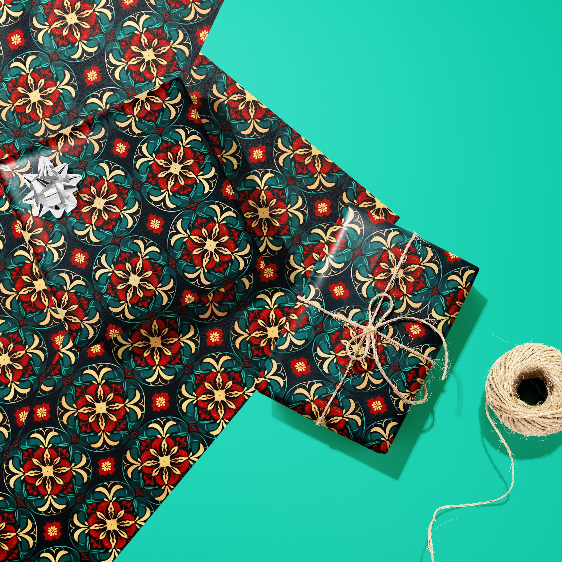 Two gifts wrapped in Arabesque Christmas paper—one with a silver bow, one tied with natural twine—rest on two matching sheets beside a twine spool on a bright teal surface.