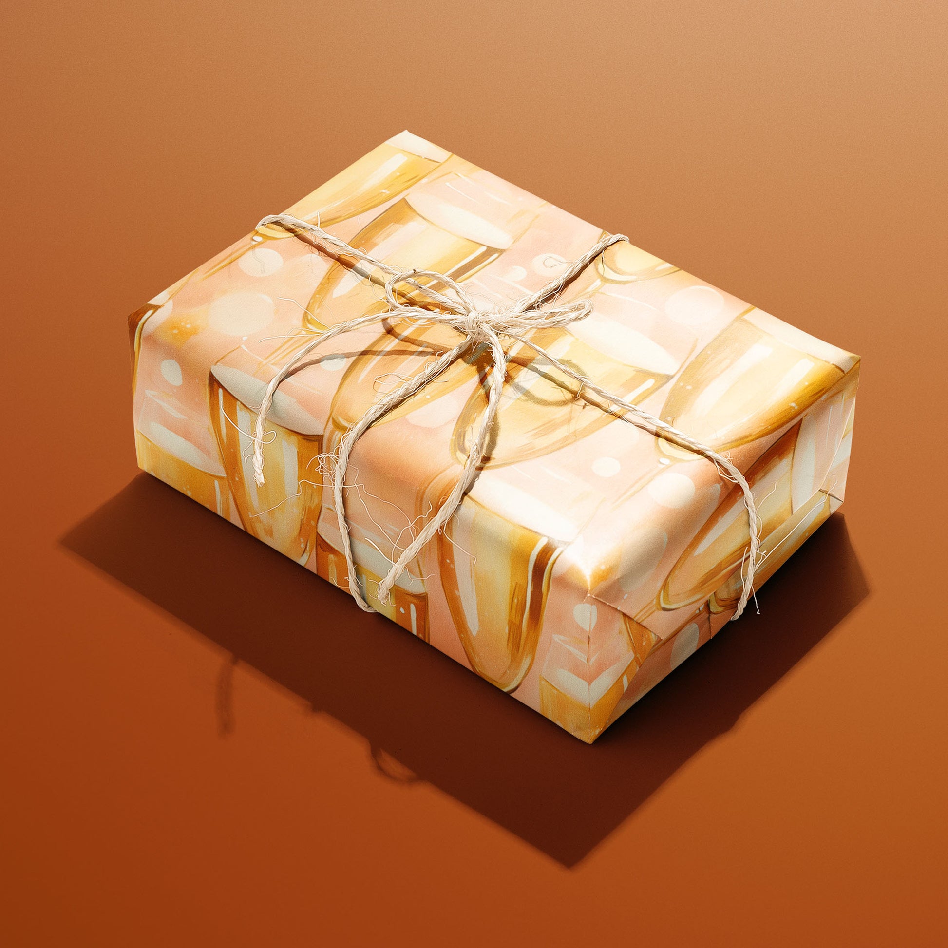 A single rectangular gift wrapped in Shimmering Sips paper—golden flutes and bubbles on peach—tied with natural twine on a copper-orange background.