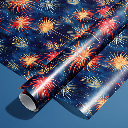 A roll of Blooming Sky wrapping paper resting on an unrolled sheet, with painterly fireworks bursts in red, gold, and white on a midnight navy print, on a steel-blue background.