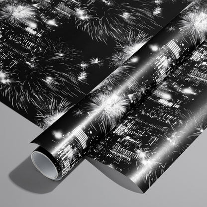 A roll on an unrolled sheet of Skyline Celebration wrapping paper with monochrome city skylines and bursting fireworks on black, set on a cool gray background.