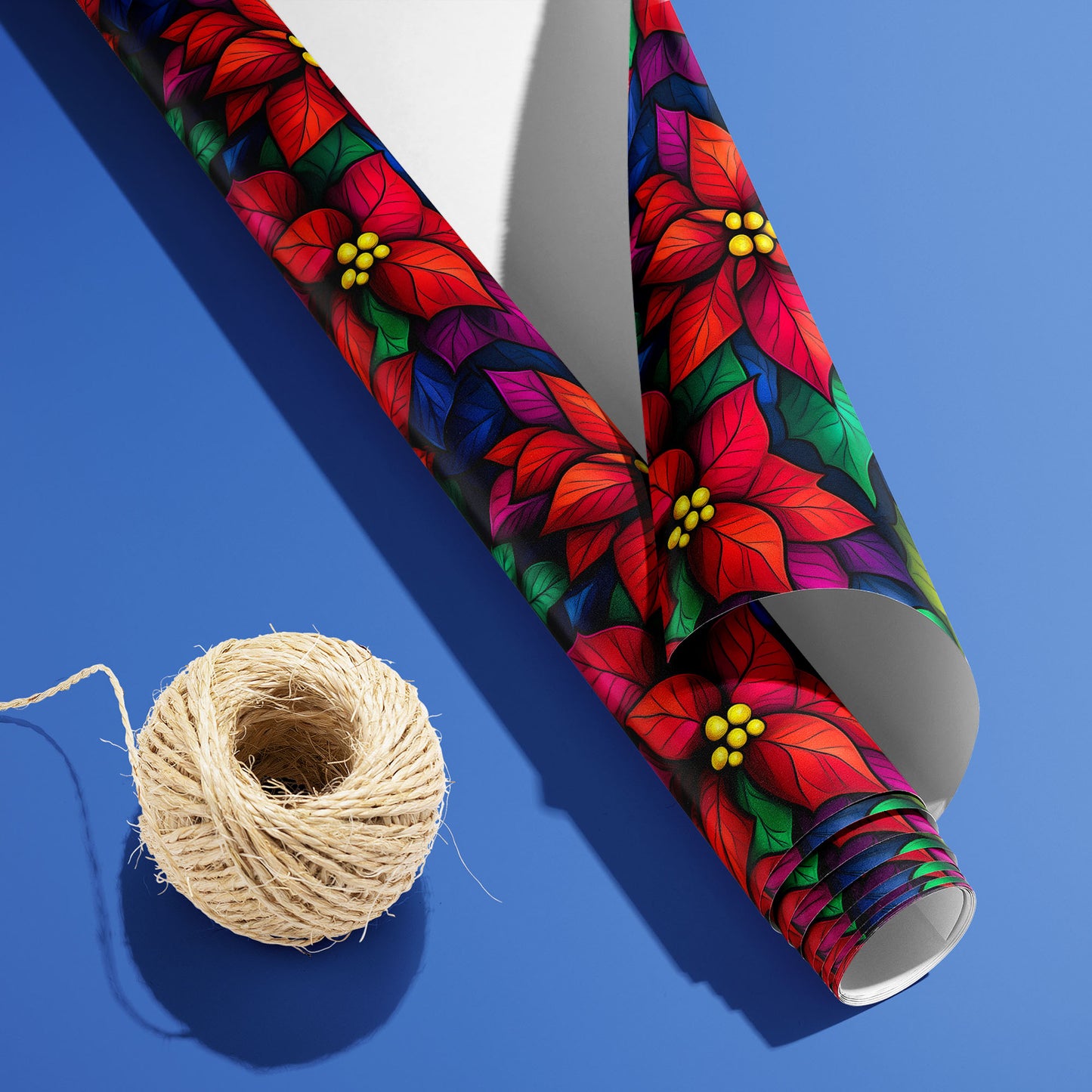 A partially unfurled roll of Bold Blooms with the white underside visible beside a spool of natural twine, displaying luminous poinsettias on black over a cobalt-blue surface.