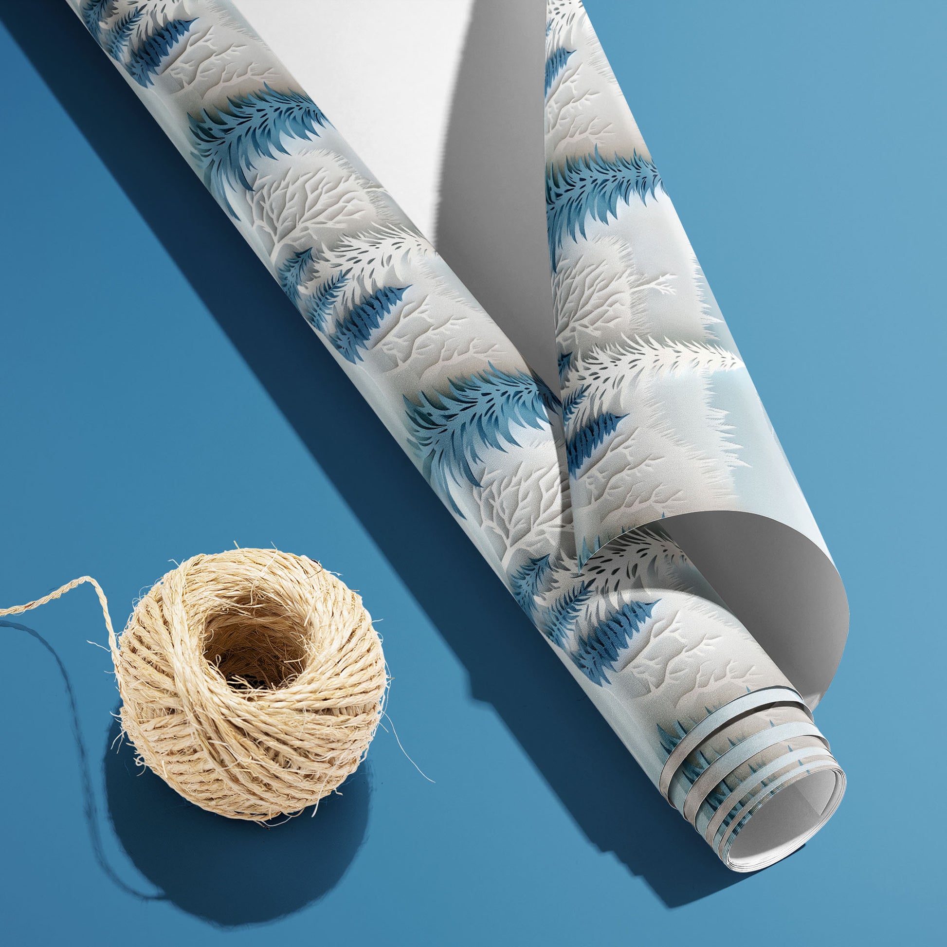 A partially unfurled Snowy Grove roll with the white underside visible beside a spool of natural twine, featuring paper-cut winter trees in cool blues on a medium blue background.