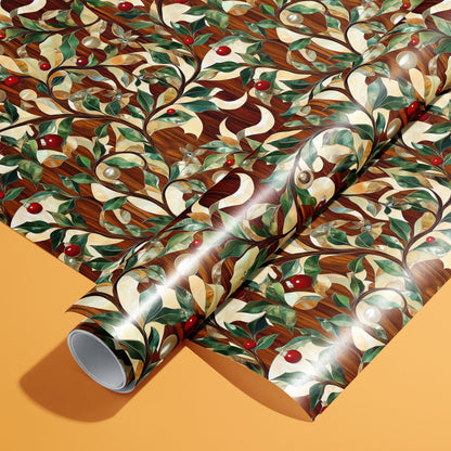 A full roll resting on an unrolled sheet of Gilded Greenery wrapping paper with gilded vines, green leaves, red berries, and pearl-like baubles over woodgrain, set on a warm orange background.
