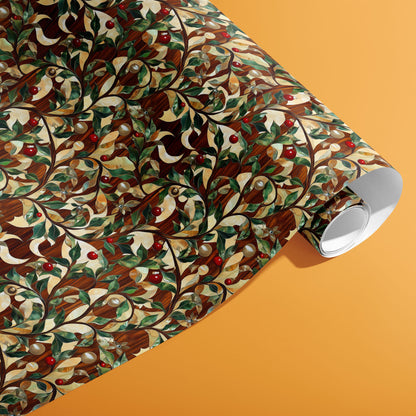 A partially unfurled roll of Gilded Greenery wrapping paper showing gold-touched vines, lush leaves, and red berries on a woodgrain pattern atop a warm orange background.