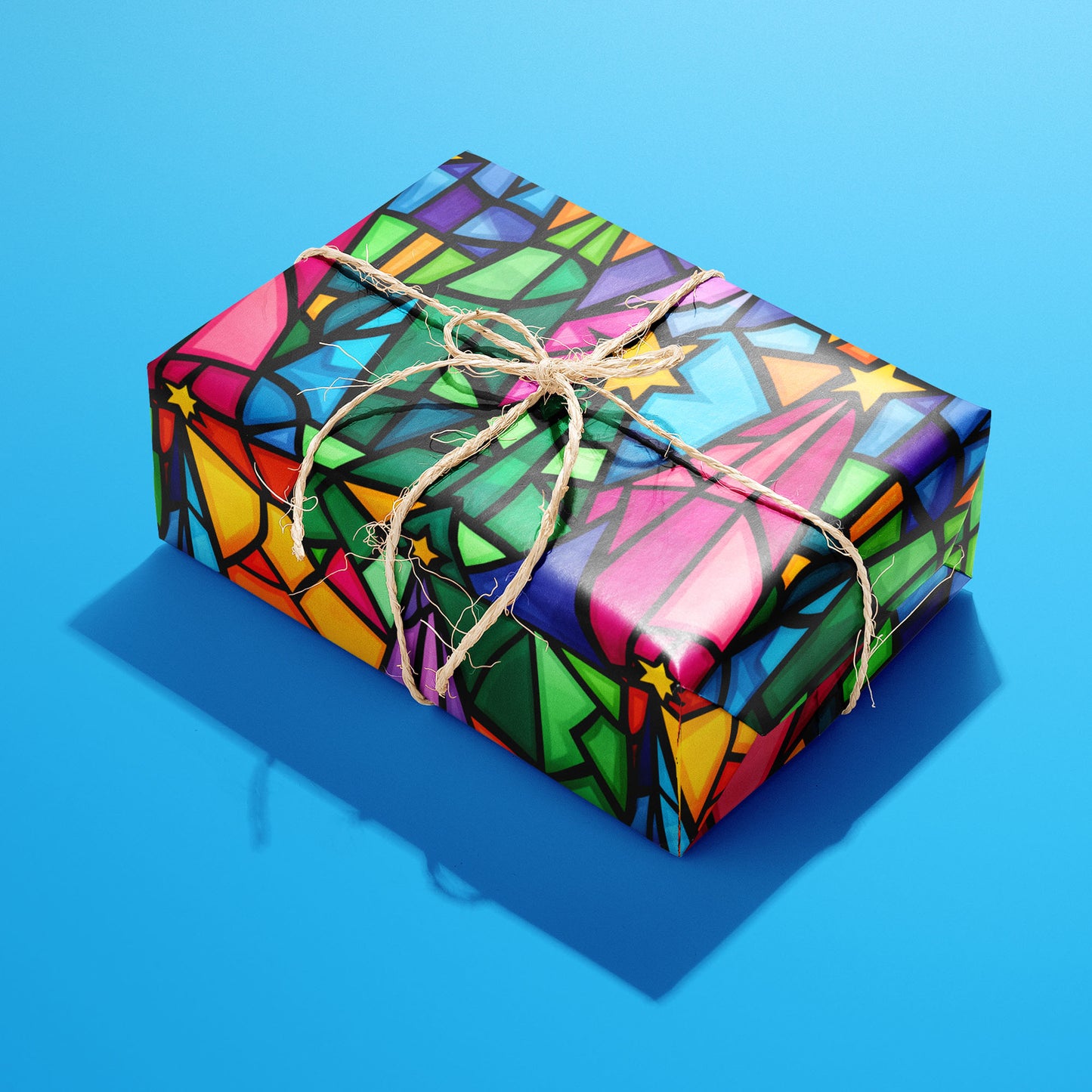 A single rectangular gift wrapped in Christmas Kaleidoscope and tied with natural twine, the stained-glass star pattern glowing in rainbow colors against a bright blue background.