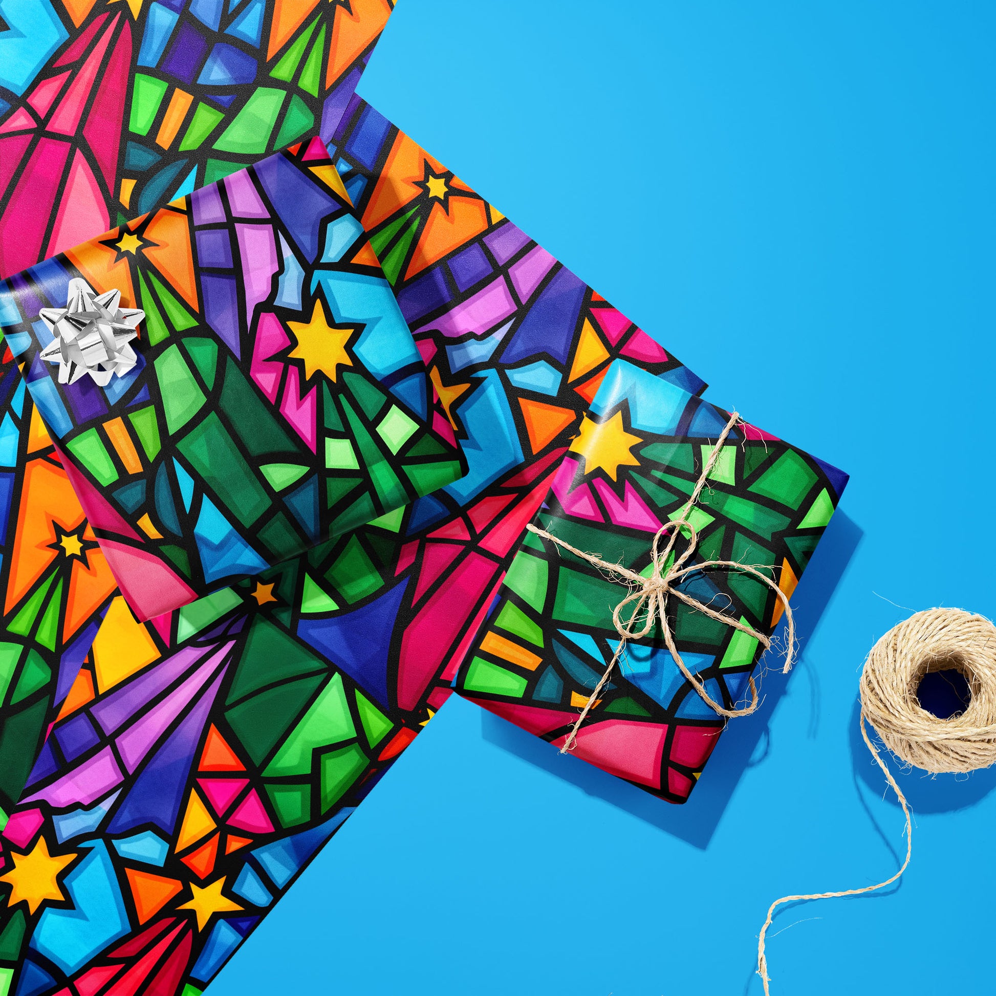 Two gifts in Christmas Kaleidoscope—one with a silver bow and one tied with natural twine—rest on two unused sheets beside a spool of twine on a bright blue background.