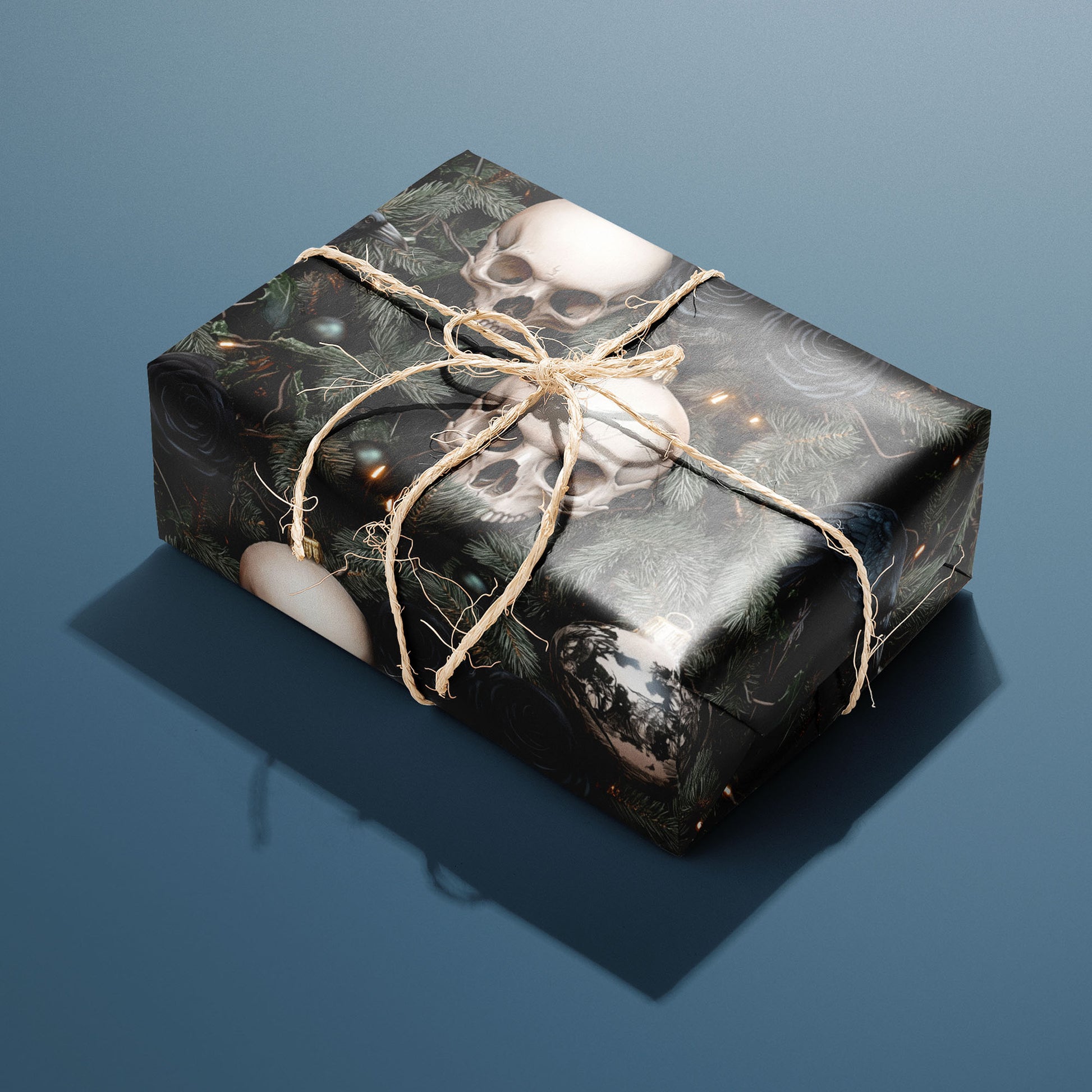 A single gift wrapped in Dark Yuletide and tied with natural twine displays skull baubles, dark roses, and pine with tiny lights on a black ground against a steel blue background.
