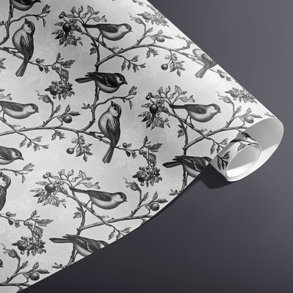 A partially unfurled roll of Winter Aviary gift wrap reveals monochrome birds and berries on a subtle damask white ground against a charcoal gray background.