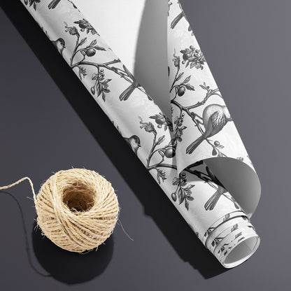 A partially unfurled roll of Winter Aviary, white underside visible beside a spool of natural twine, shows engraved birds on branching berries over a soft white damask ground on charcoal gray.