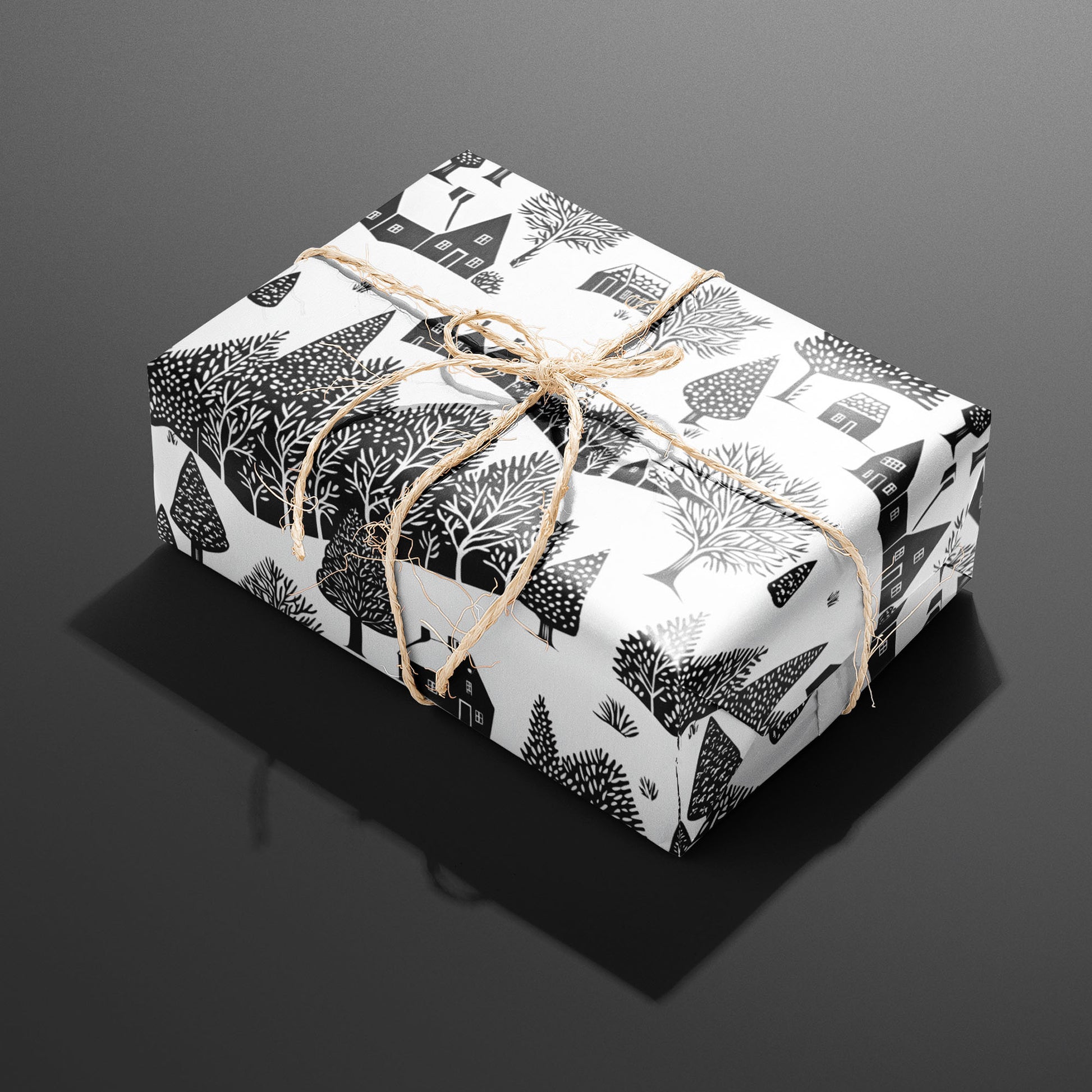 A single rectangular gift wrapped in Snowbound Village and tied with natural twine, the black cottages and snow-laden trees crisp on a white ground over dark gray.