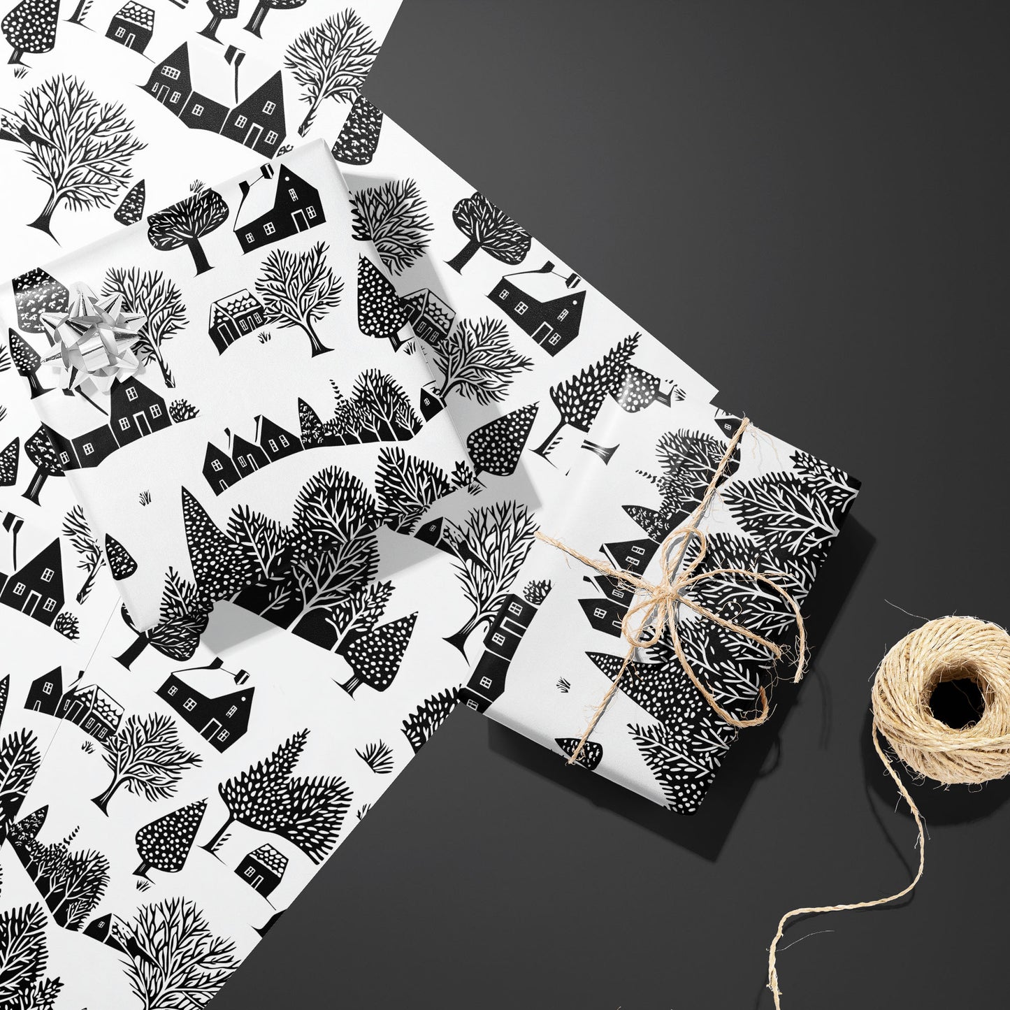 Two gifts in Snowbound Village—one with a silver bow and one tied with twine—rest on two unused sheets beside a twine spool on dark gray, with black cottages and snowy trees.