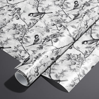 A roll on an unrolled sheet of Winter Orchard wrapping paper with etched songbirds on fruiting branches over a frosty gray ground, set on a dark charcoal background.