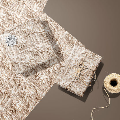 Two gifts in Winter Weave—one with a silver bow, one tied with twine—rest on two unused sheets beside a twine spool on a warm brown surface, showing a knit snowflake pattern.