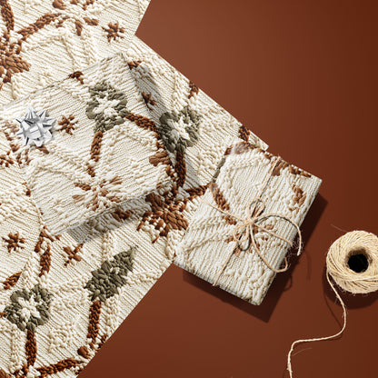 Two Festive Fibers gifts—one with a silver bow and one tied with twine—rest on two unused sheets beside a twine spool on a terracotta surface, showing knit stars and trees.