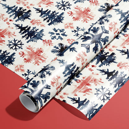 A roll of Scandinavian Snowflakes wrapping paper resting on a matching unrolled sheet, featuring navy and red snowflakes and trees on cream over a red background.