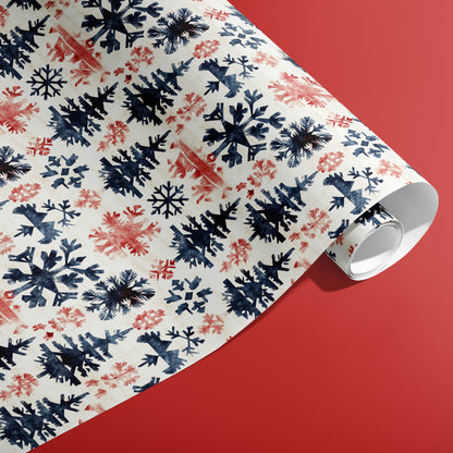 A partially unfurled roll of Scandinavian Snowflakes gift wrap showing painterly navy and red snowflakes and fir trees on cream paper against a warm red surface.