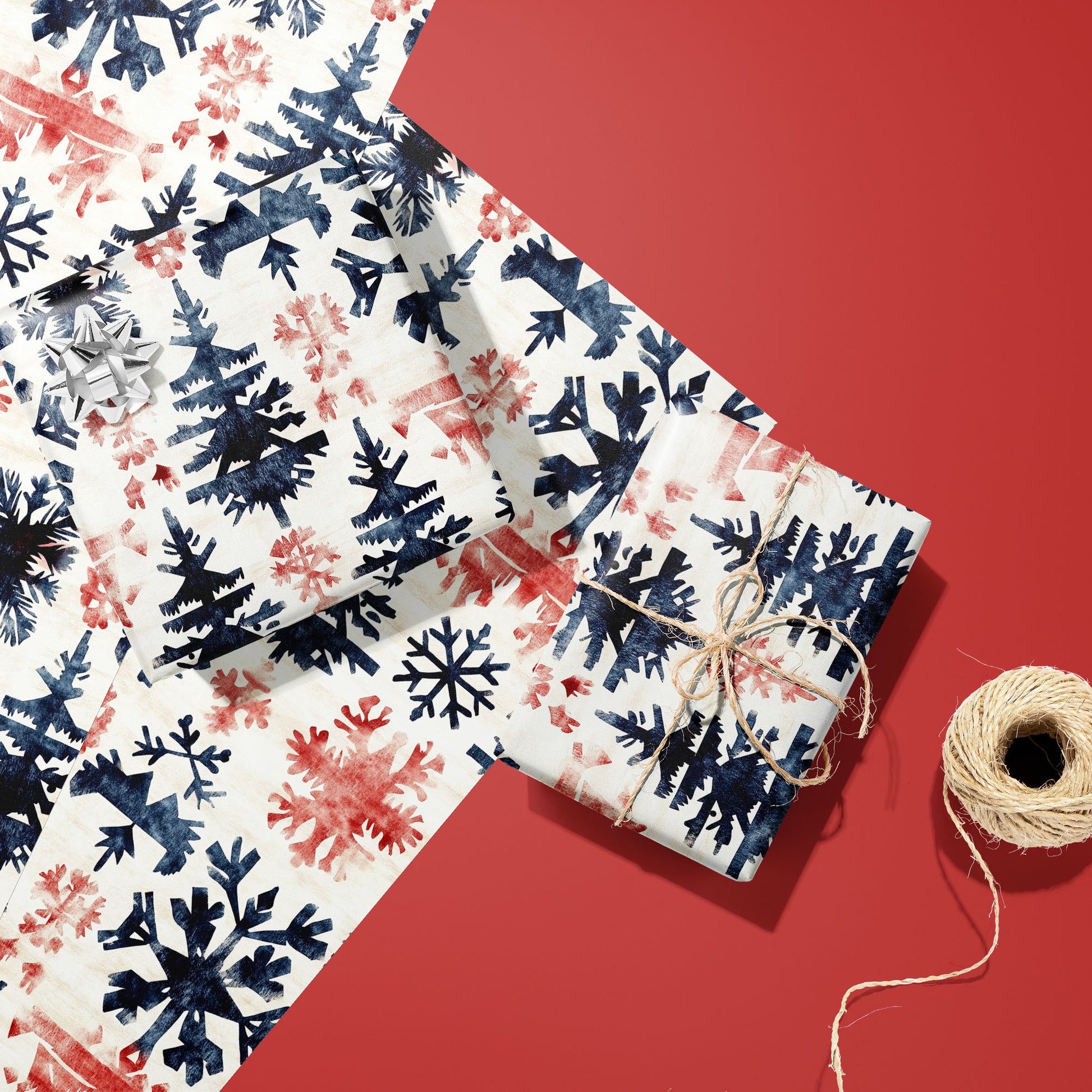 Two gifts wrapped in Scandinavian Snowflakes paper, one with a silver bow and one tied with twine, resting on extra sheets beside a spool of twine on a red surface.