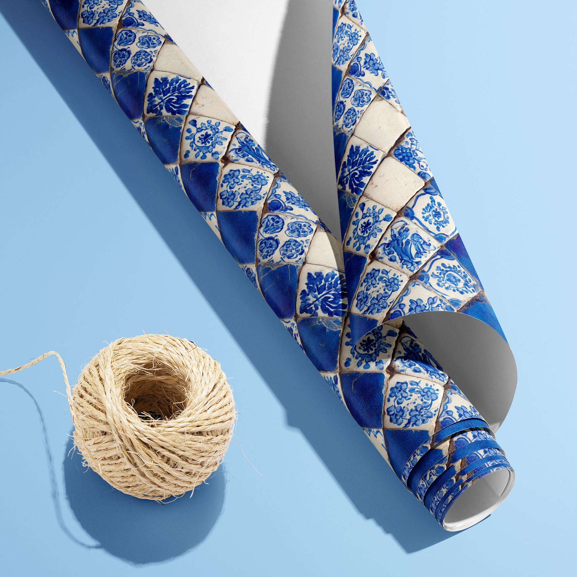 A partially unfurled roll of Ionian Inks wrapping paper with the white underside showing beside a spool of natural twine on a soft blue backdrop, patterned with Delft-style tiles.