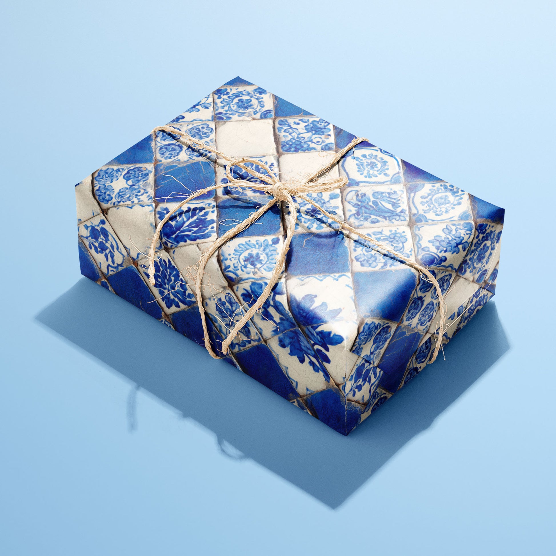 A rectangular gift wrapped in Ionian Inks paper, the blue-and-white tile pattern tied with a natural twine bow, set on a light blue background.