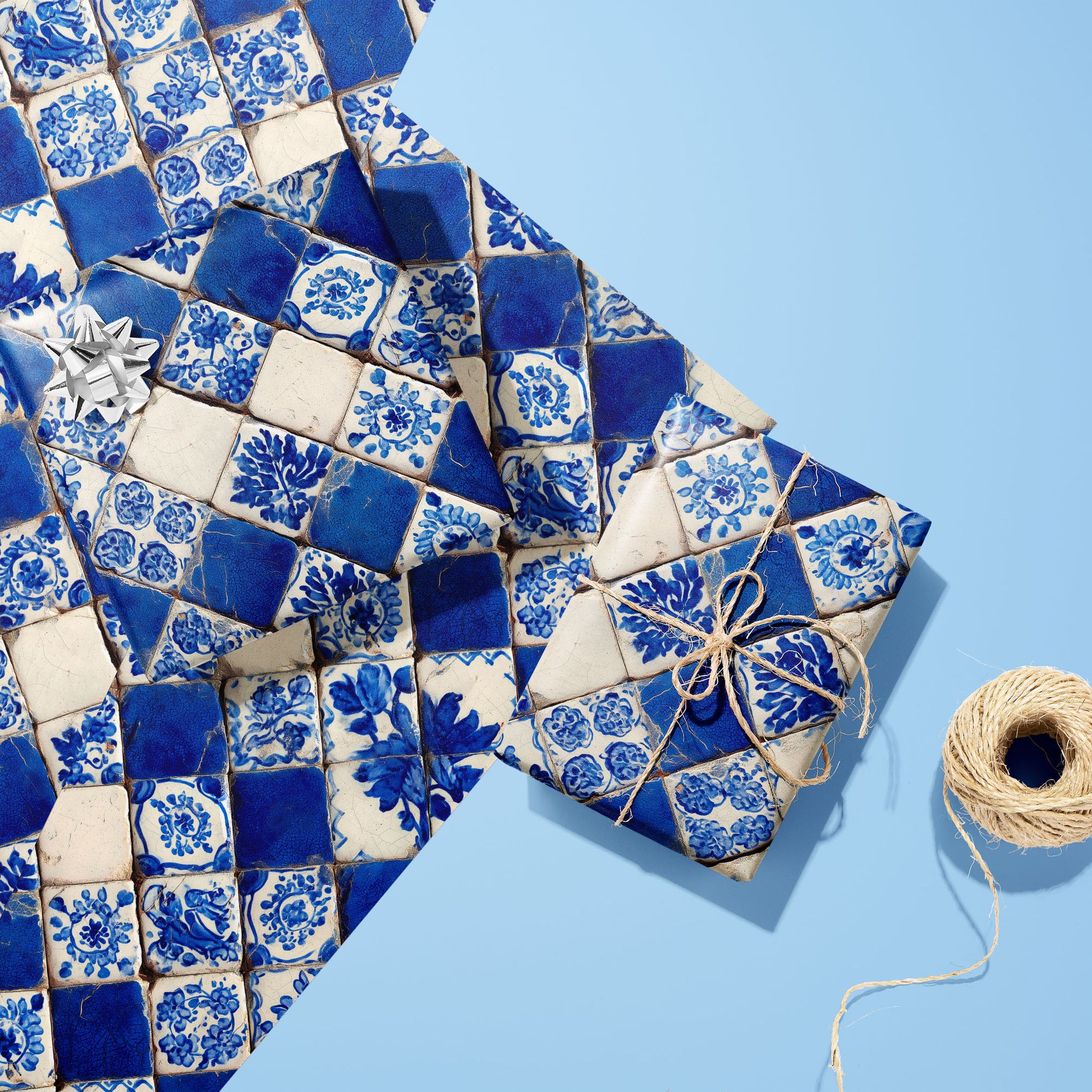 Two gifts wrapped in Ionian Inks paper—one with a silver bow, one tied in twine—rest on two unused sheets beside a twine spool on a pale blue surface.