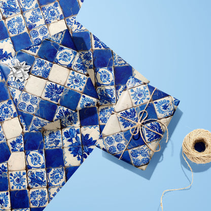 Two gifts wrapped in Ionian Inks paper—one with a silver bow, one tied in twine—rest on two unused sheets beside a twine spool on a pale blue surface.