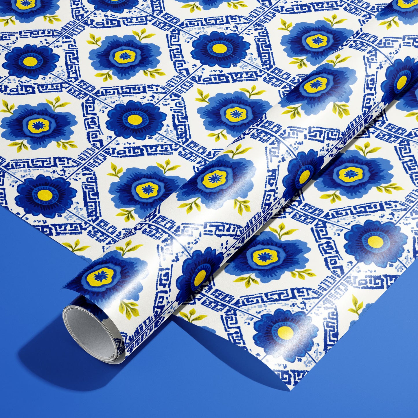 A roll of Santorini Floriate wrapping paper resting on an unrolled sheet, featuring cobalt floral tile motifs with Greek-key lines on white, set against a bold blue background.
