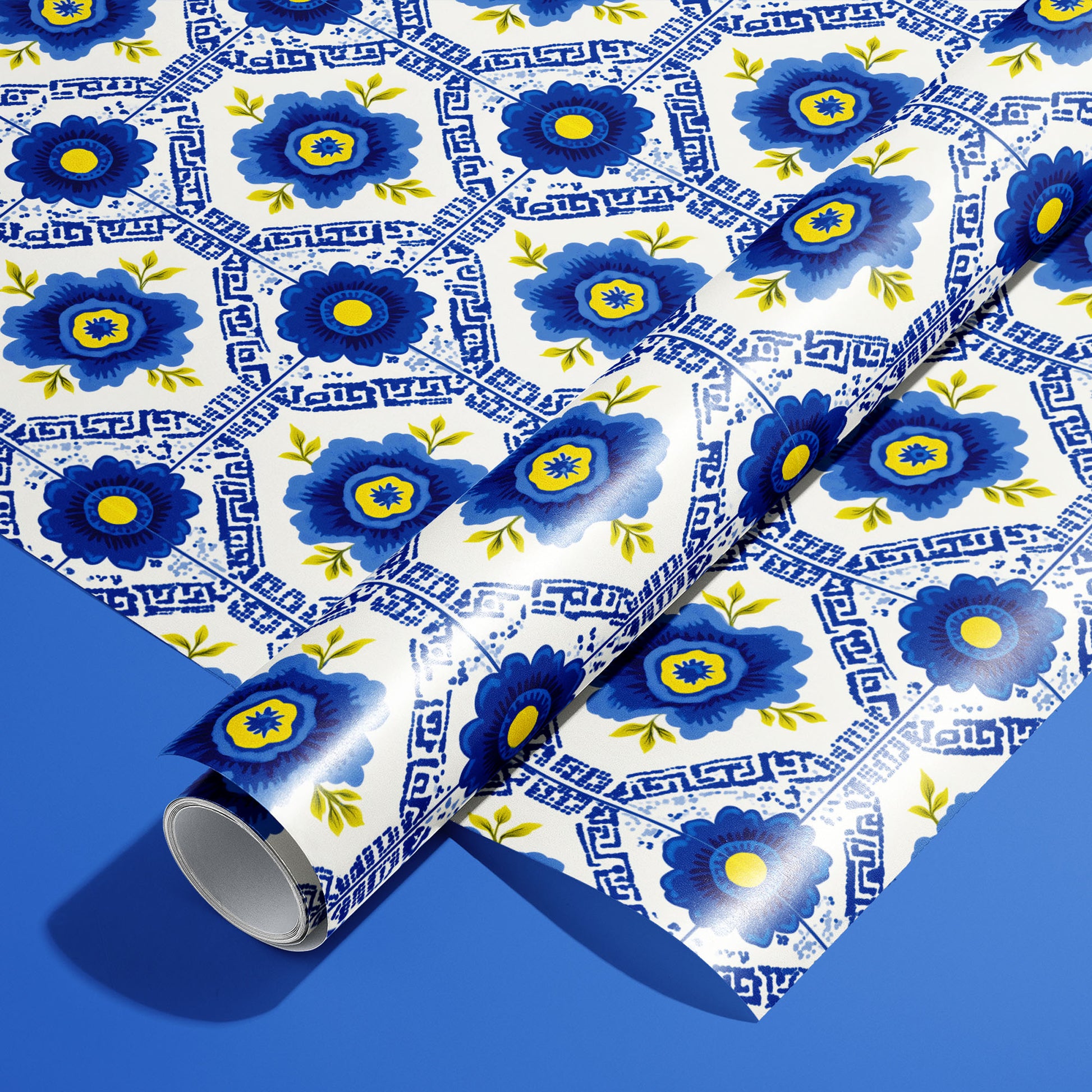 A roll of Santorini Floriate wrapping paper resting on an unrolled sheet, featuring cobalt floral tile motifs with Greek-key lines on white, set against a bold blue background.