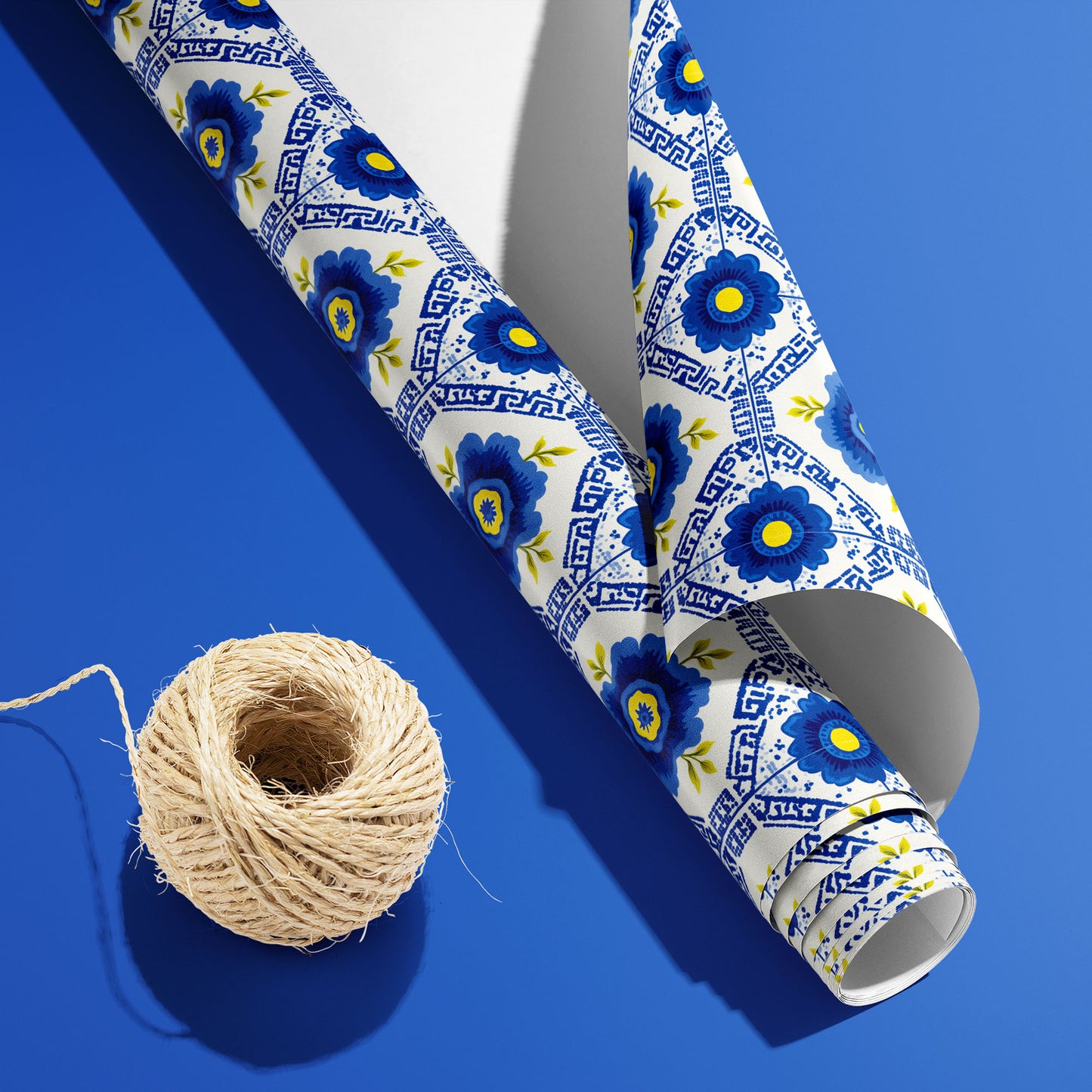 A partially unfurled roll of Santorini Floriate with the white underside showing beside a spool of natural twine, patterned with blue florals and Greek-key tiles on a blue surface.