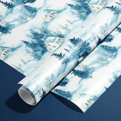 A roll of Cozy Cottage wrapping paper resting on a matching unrolled sheet, featuring watercolor snow-covered cottages and pine trees on soft blue-white against a navy background.