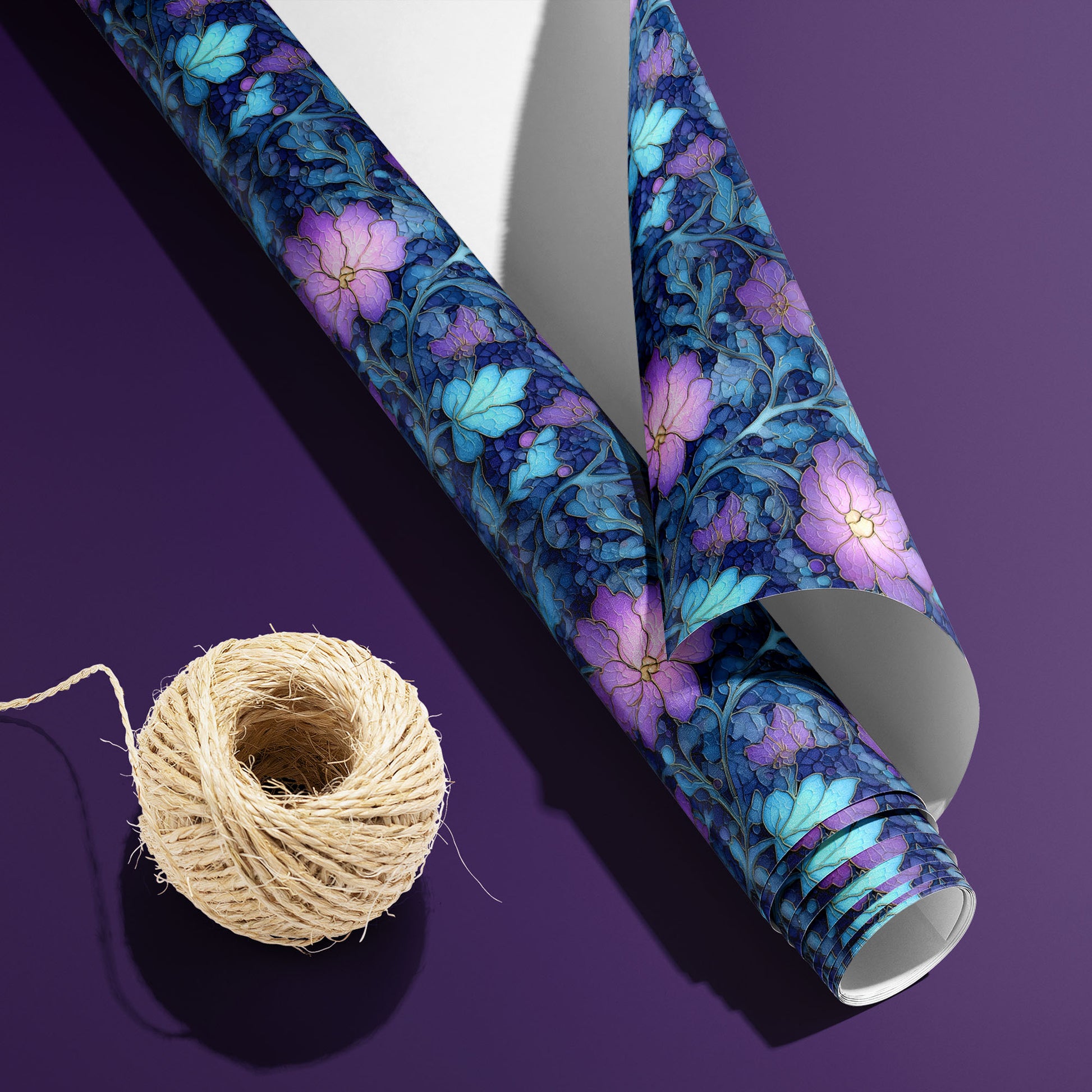 A partially unfurled roll of Lunar Hellebore with its white underside visible beside a spool of natural twine, the violet-and-teal floral print on midnight blue, on a purple surface.