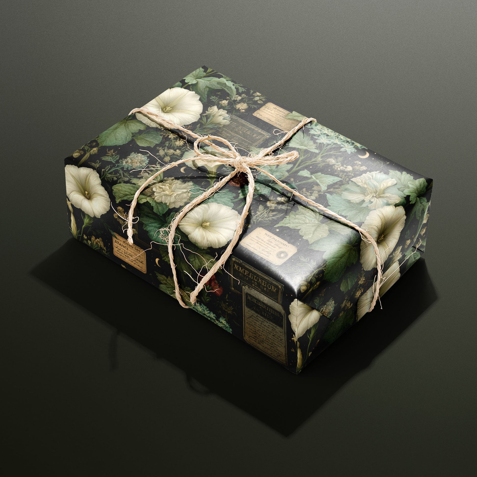 A single gift wrapped in Vesper Botanica and tied with natural twine, displaying cream flowers, leafy botanicals, and antique labels on black, on a dark olive green surface.